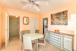A dining area with a glass table and chairs, a white ceiling fan, a cabinet with drawers, and a painting on the wall.