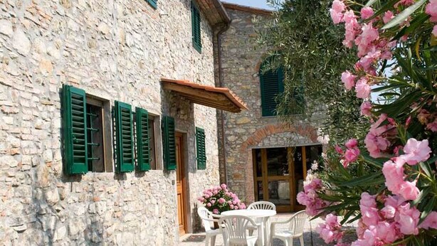 Ein Steinhaus mit grünen Fensterläden, einer Terrasse mit weißen Stühlen und rosa Blumen.