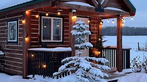 A wooden cabin with a porch, a snow-covered tree, and string lights. - Whispering Winds Cabin - cozy double loft cabin (Pickardville)