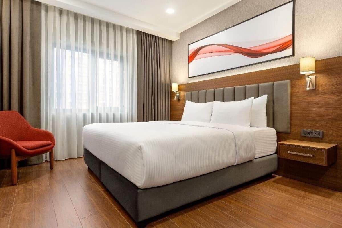 A hotel room with a large bed, a red chair, a wooden nightstand, and a wall-mounted lamp.