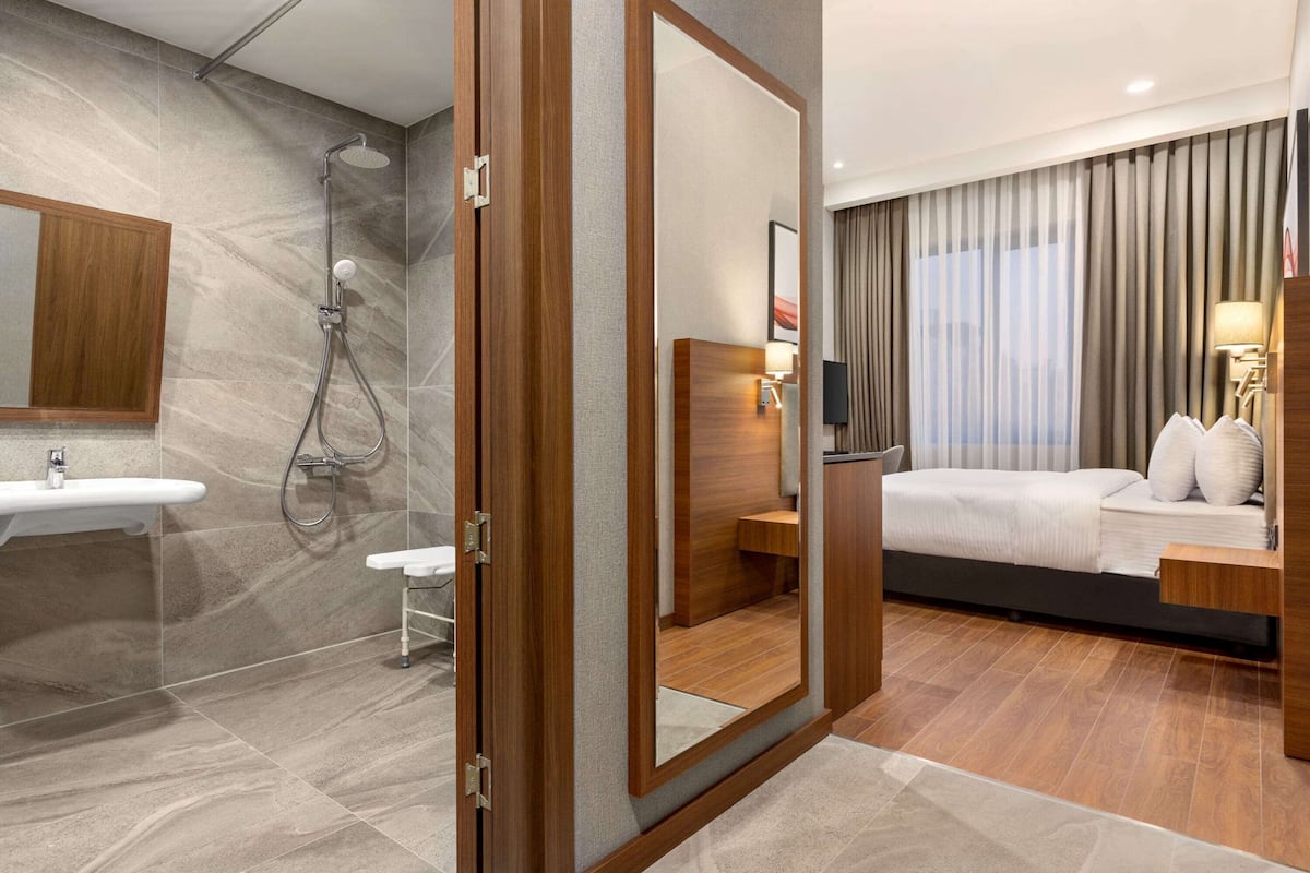A modern hotel room with a wooden bed, a bedside table, a nightstand, a mirror, a bathroom with a sink and shower.