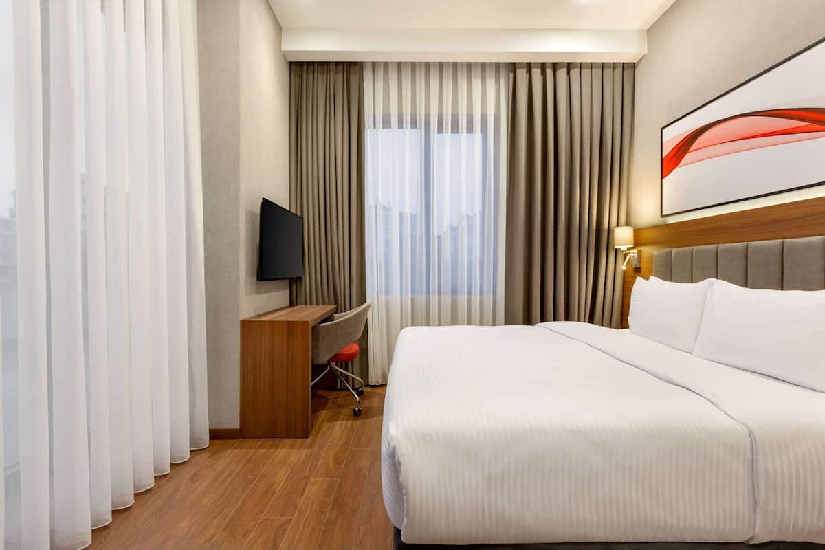 A hotel room with a large bed, a desk with a chair, a TV mounted on the wall, and a window with curtains.
