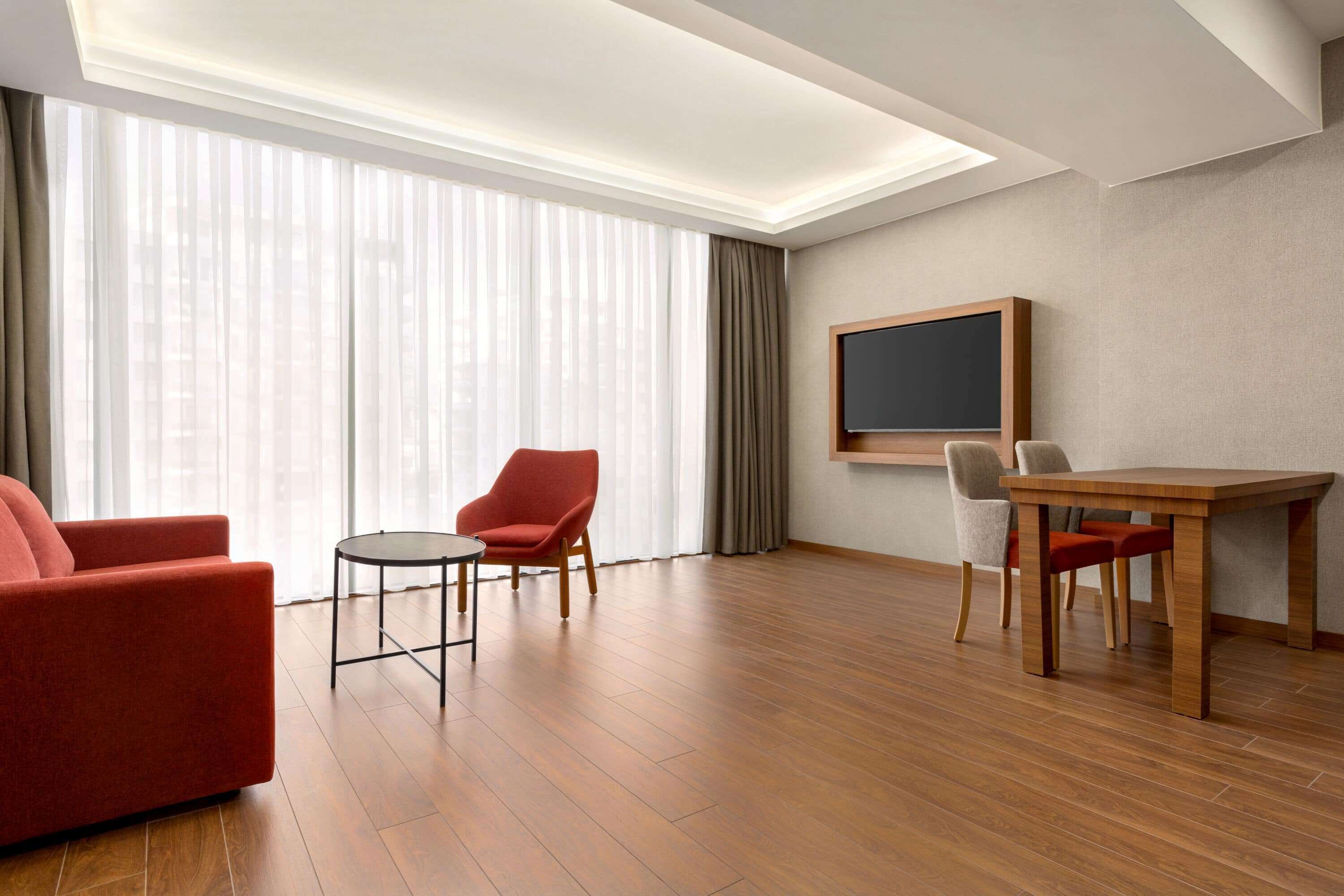 Una habitación de hotel moderna con suelo de madera, un sofá rojo, una pequeña mesa redonda, un sillón rojo, una mesa de comedor de madera con sillas y un gran ventanal con cortinas traslúcidas.