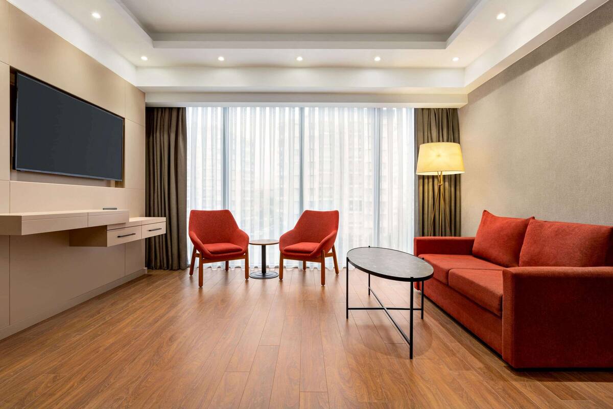 A modern hotel room with a flat-screen TV, two red armchairs, a red sofa, a small round table, and a large window with sheer curtains.