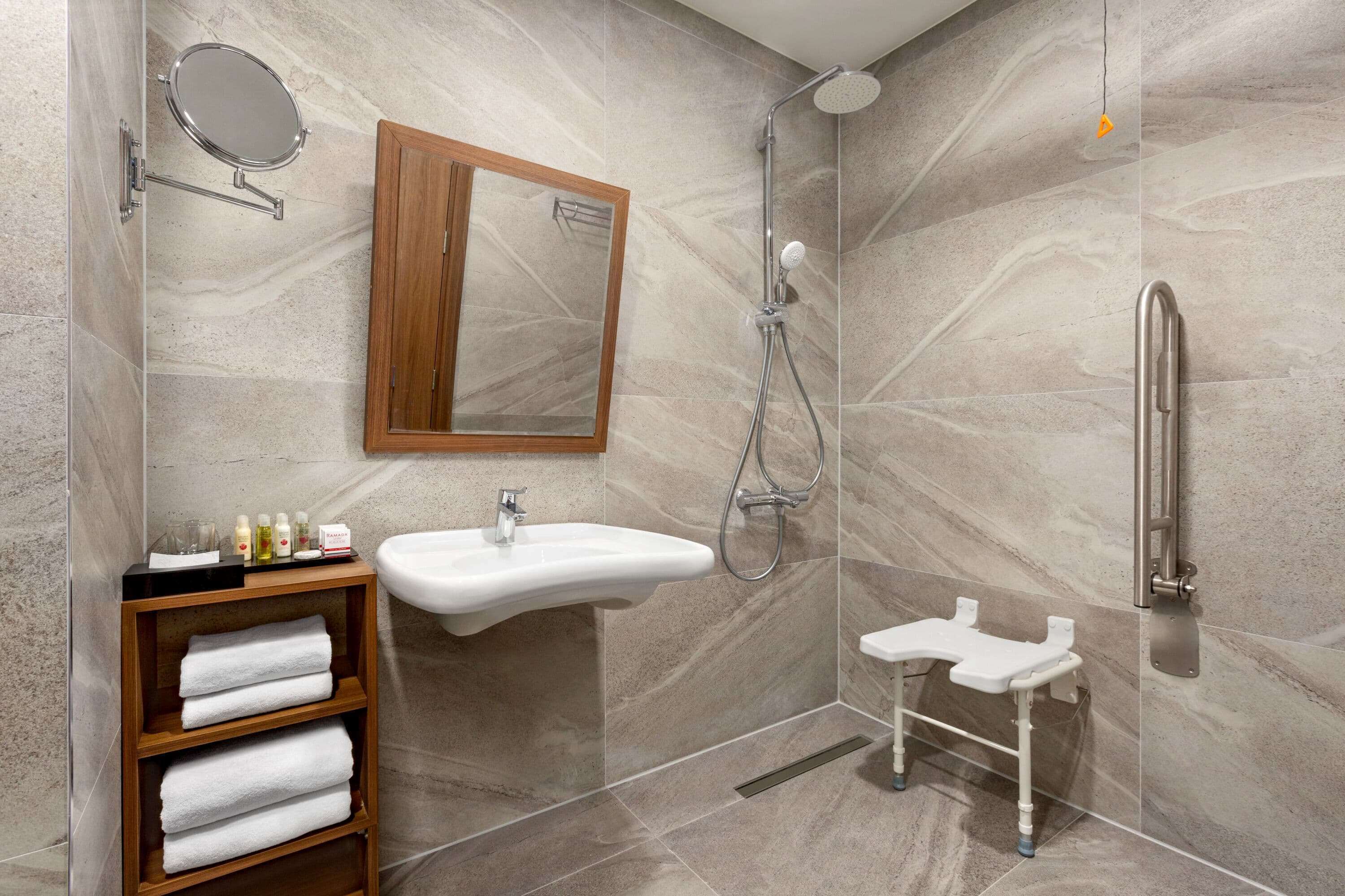 Un baño moderno con pared de mármol, tocador de madera con toallas, lavamanos, espejo, ducha con cabezal de ducha manual y barra de apoyo.