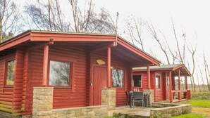 A log cabin with a porch, stone foundation, and a small outdoor seating area. - Ingram (Morpeth)