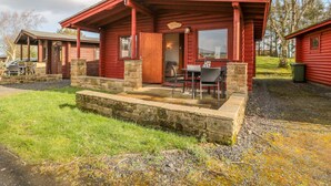 A red log cabin with a stone foundation, a porch with chairs, and a table. - Ingram (Morpeth)