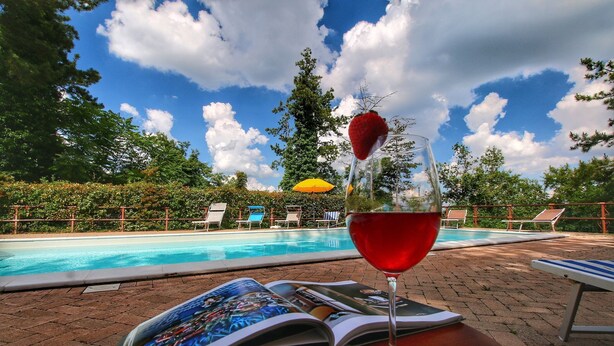 A poolside setting with a glass of red wine, a book, and lounge chairs.