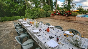 An outdoor dining area with a long table set for a meal, surrounded by greenery and a pool in the background.
