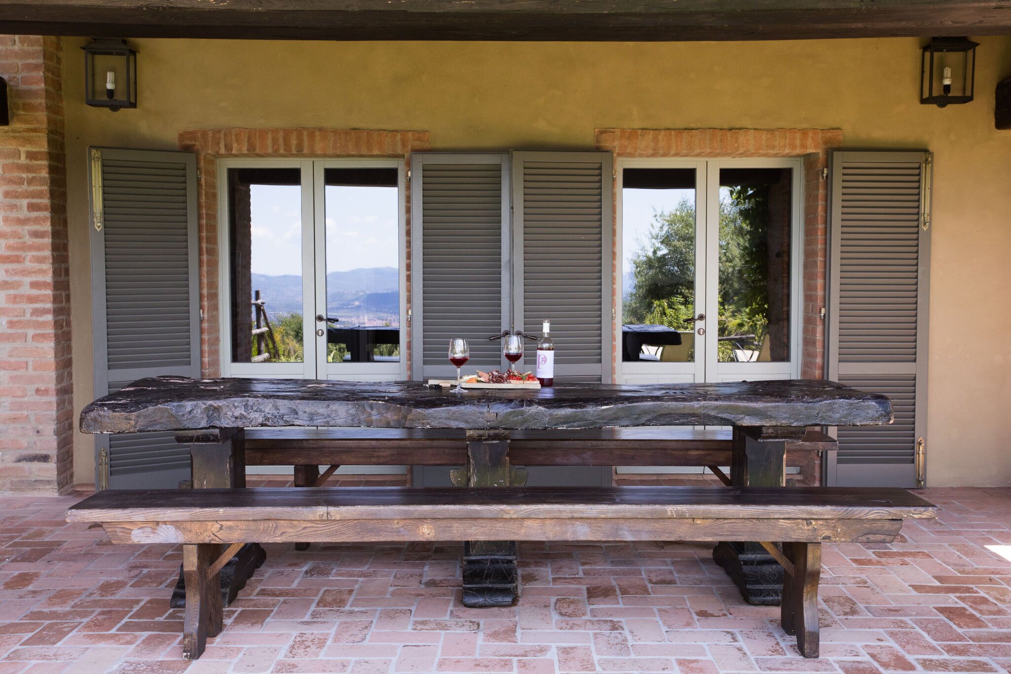 A rustic outdoor dining area with a long wooden table and benches, flanked by shuttered windows with a scenic view.
