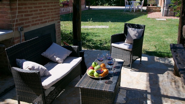 An outdoor seating area with wicker furniture, a table with fruit and wine glasses, and a brick structure in the background.