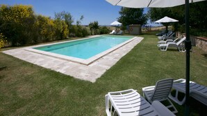 A swimming pool with a paved deck, surrounded by grass and trees. There are lounge chairs and umbrellas for sunbathing.