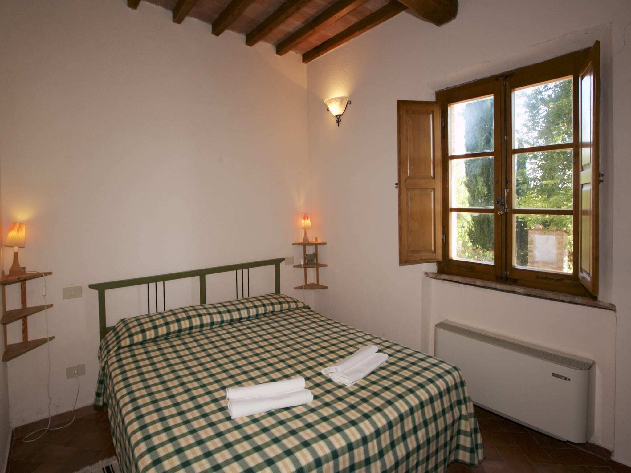 Une chambre à coucher avec un lit, deux lampes de chevet, une fenêtre donnant sur un espace verdoyant et un radiateur.
