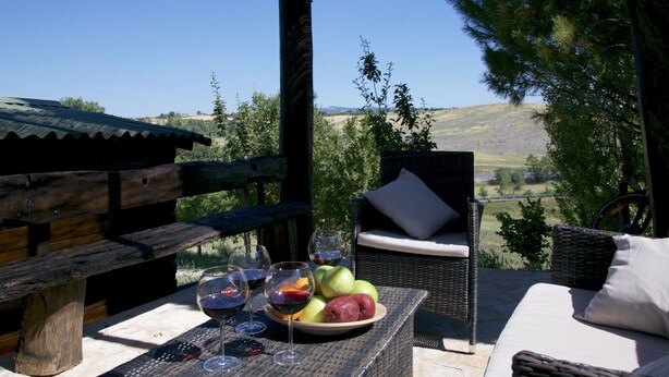 Un espacio al aire libre con mobiliario de mimbre, una mesa con una fuente de fruta y copas de vino, y una vista panorámica de colinas y árboles.
