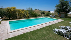 Una piscina rectangular con agua azul cristalina, rodeada por un borde de piedra. Hay sillas de descanso blancas y un flotador salvavidas cerca de la piscina.