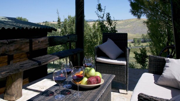 Una terraza de madera con una silla de mimbre y una mesa dispuesta con copas de vino y fruta.
