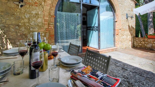 Un espace extérieur aménagé pour les repas, avec un mur en pierre, une porte vitrée donnant sur un patio couvert, et une table dressée avec des assiettes, des verres et des bouteilles de vin.