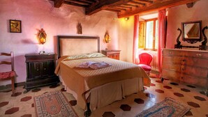 Un dormitorio con techo de madera, una cama con colcha color beige, un aparador de madera, una silla y una ventana con cortinas rojas.