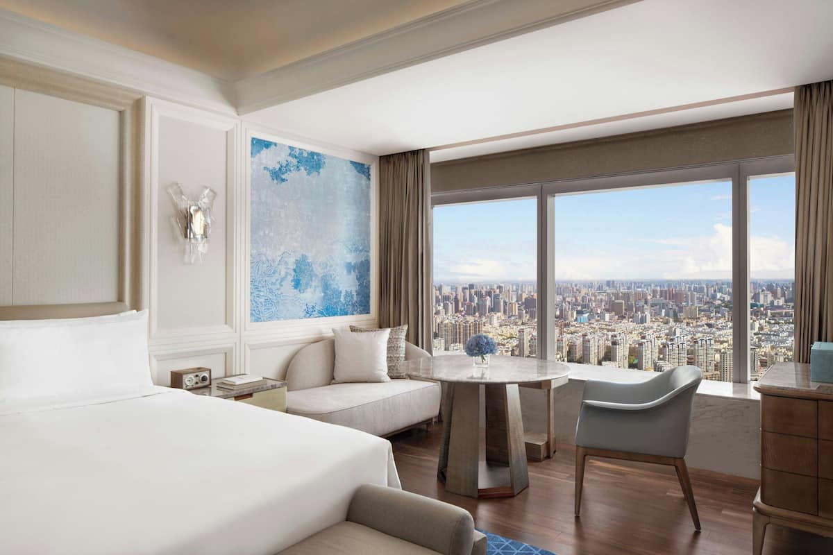 A hotel room with a large bed, a sofa, a dining table, and a chair. There is a large window with a city view.
