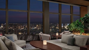 Une chambre d’hôtel moderne offrant une vue imprenable sur la ville grâce à de grandes fenêtres du sol au plafond, un coin salon confortable et une plante en pot.