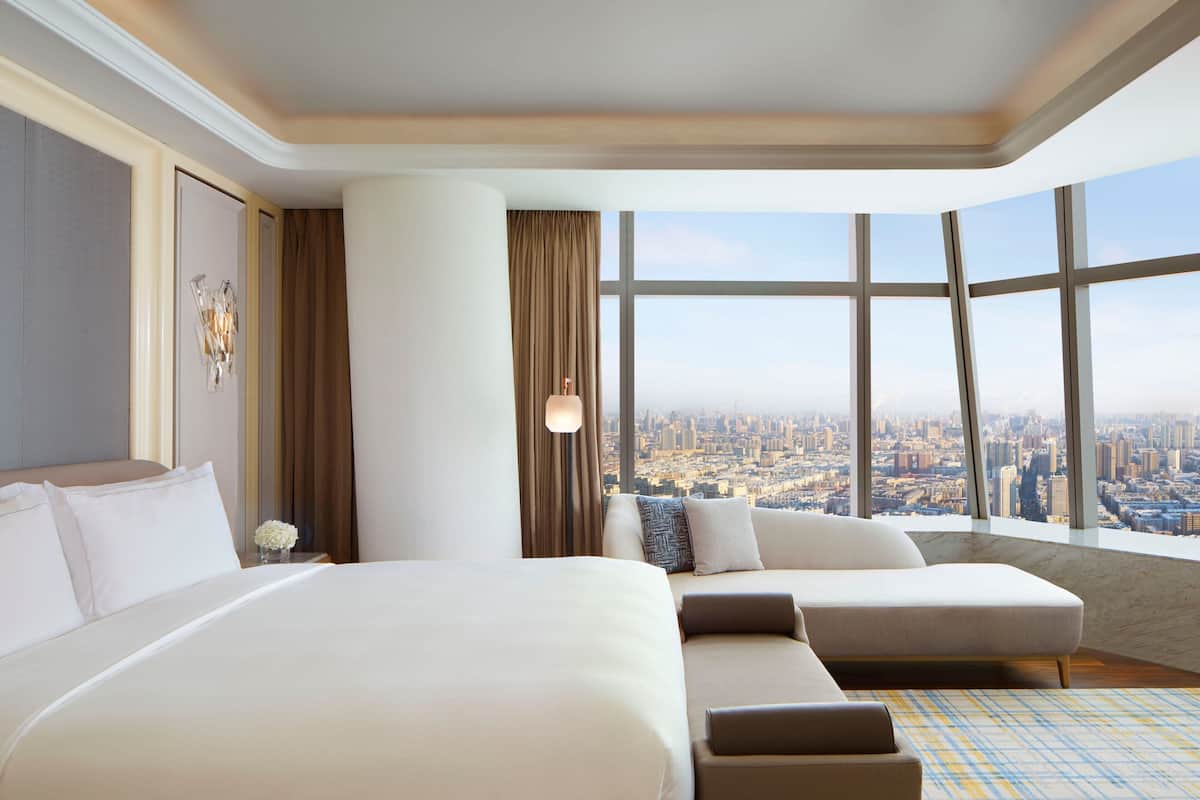 A hotel room with a large bed, a sofa, and a city view through large windows.