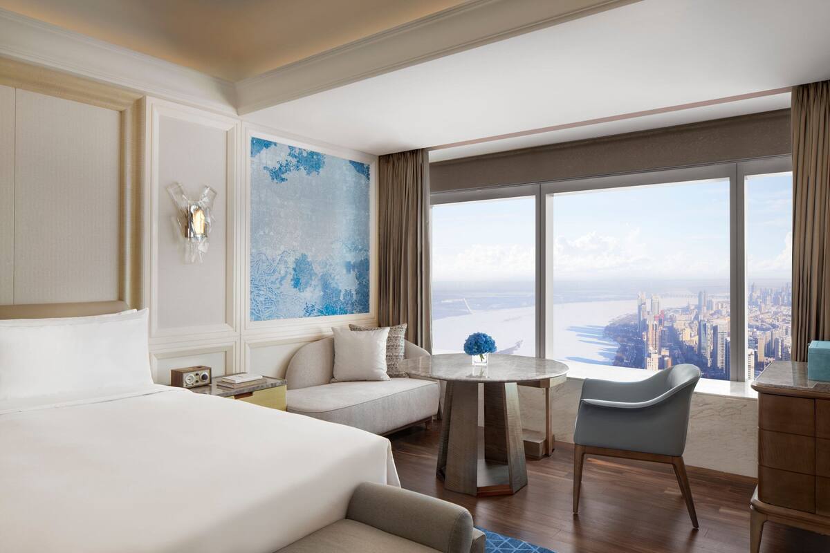 A hotel room with a large bed, a sofa, a dining table, and a view of a cityscape.
