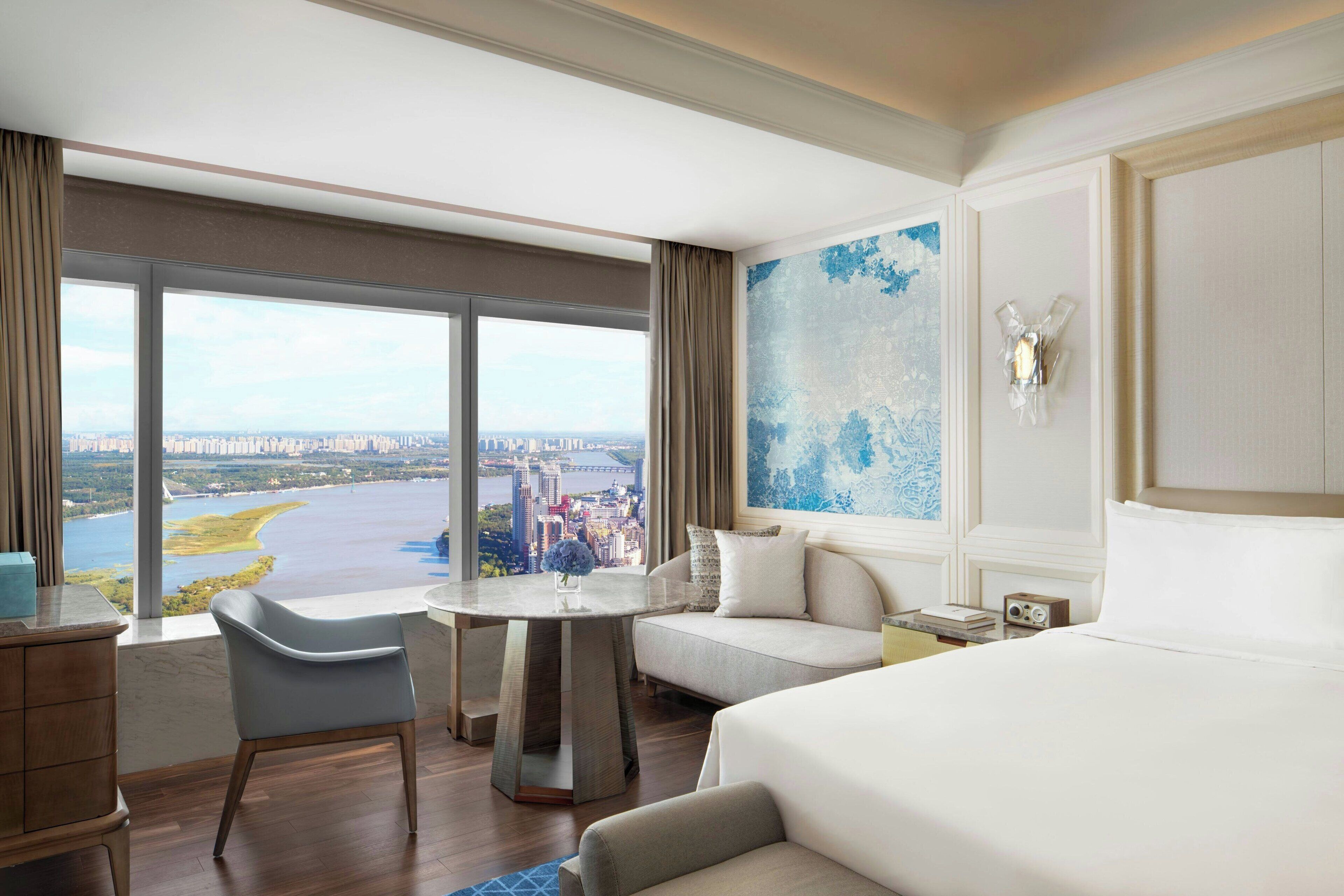 A hotel room with a large window offering a city view, a round table, a sofa, and a bed.