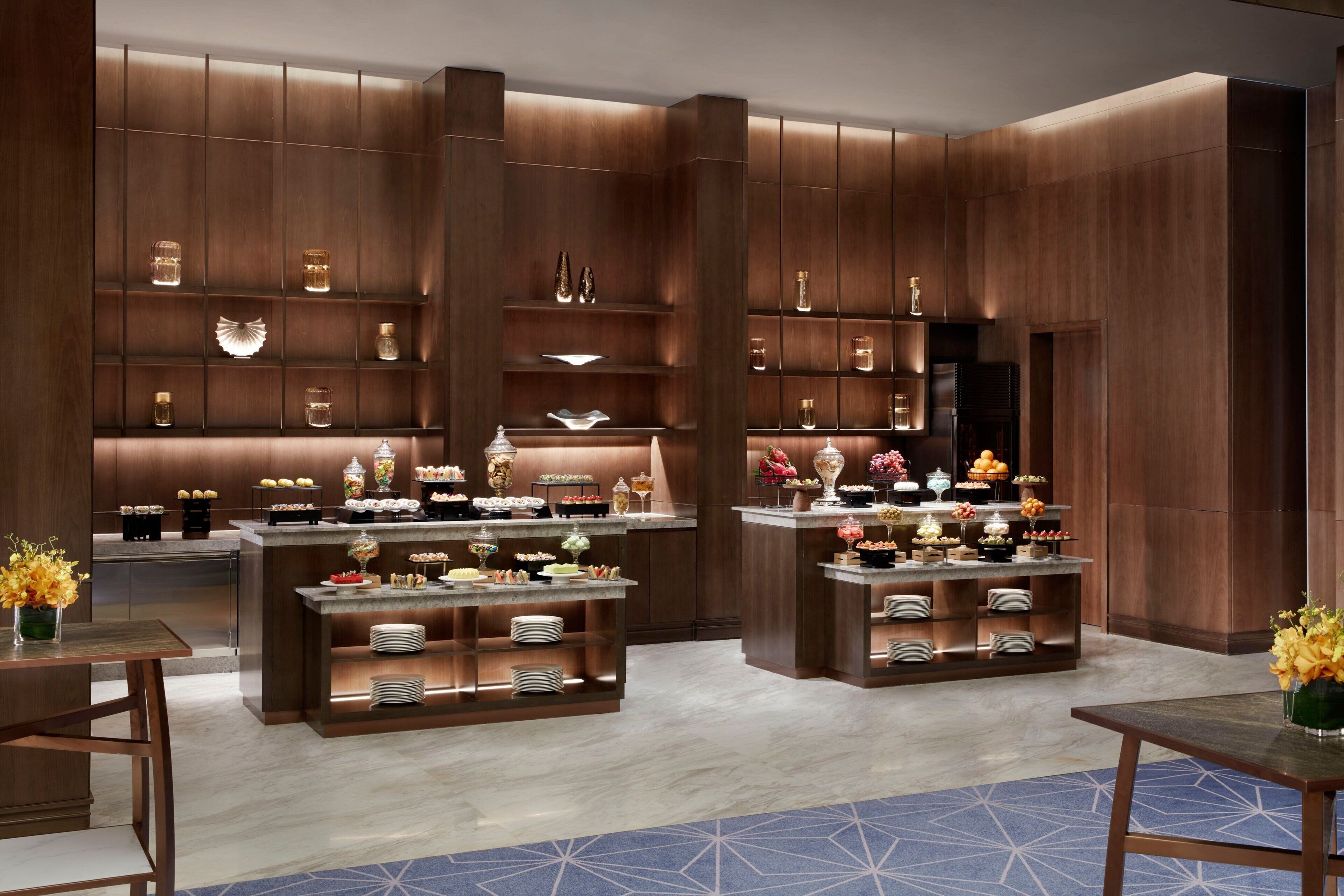 A modern hotel lobby with wooden paneling, a central reception desk, and a buffet area with various food items and decorative objects.