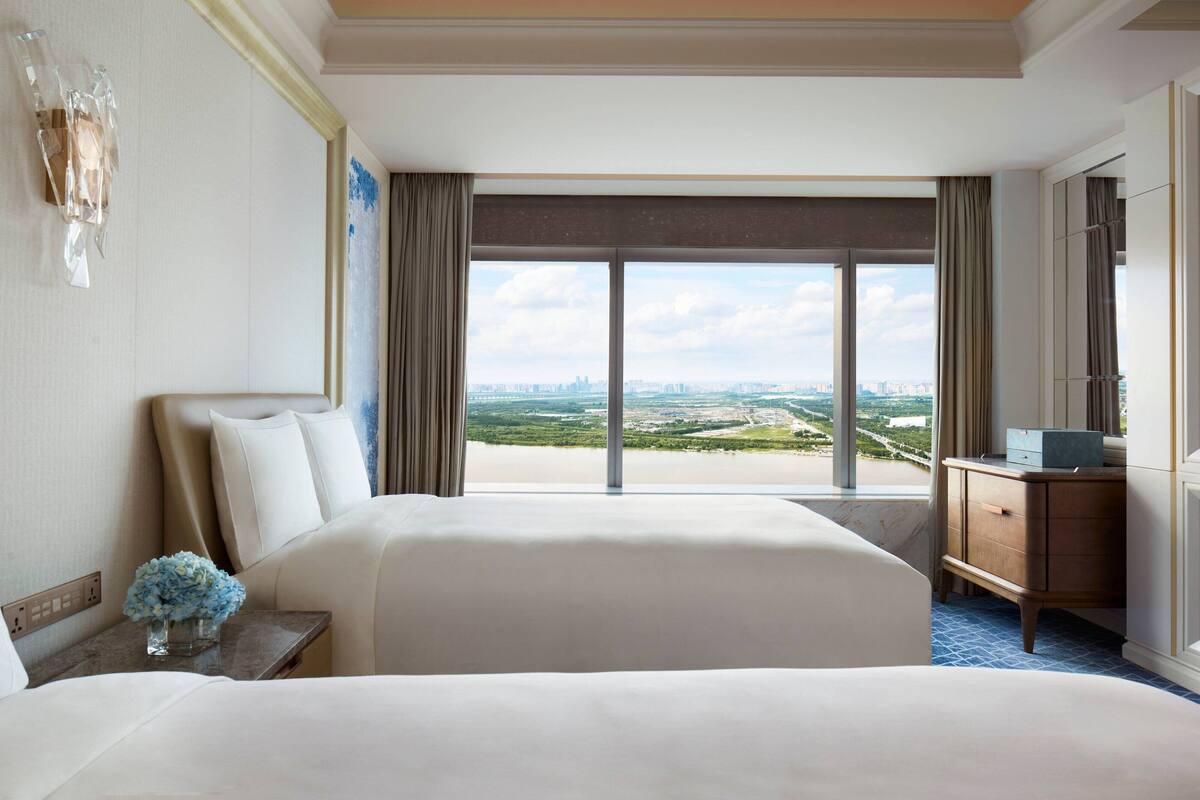 A hotel room with two beds, a nightstand with a vase of flowers, a dresser, and a large window offering a city view.