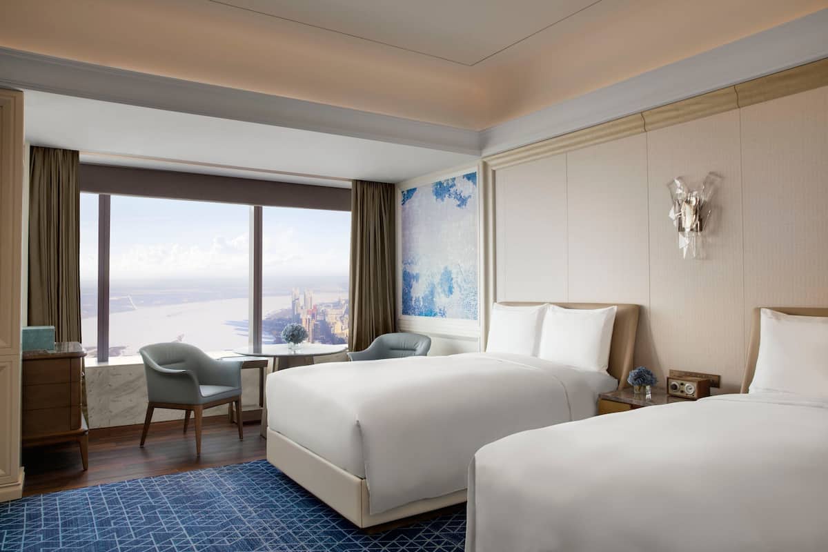 A hotel room with two beds, a desk, a chair, and a view of a cityscape.