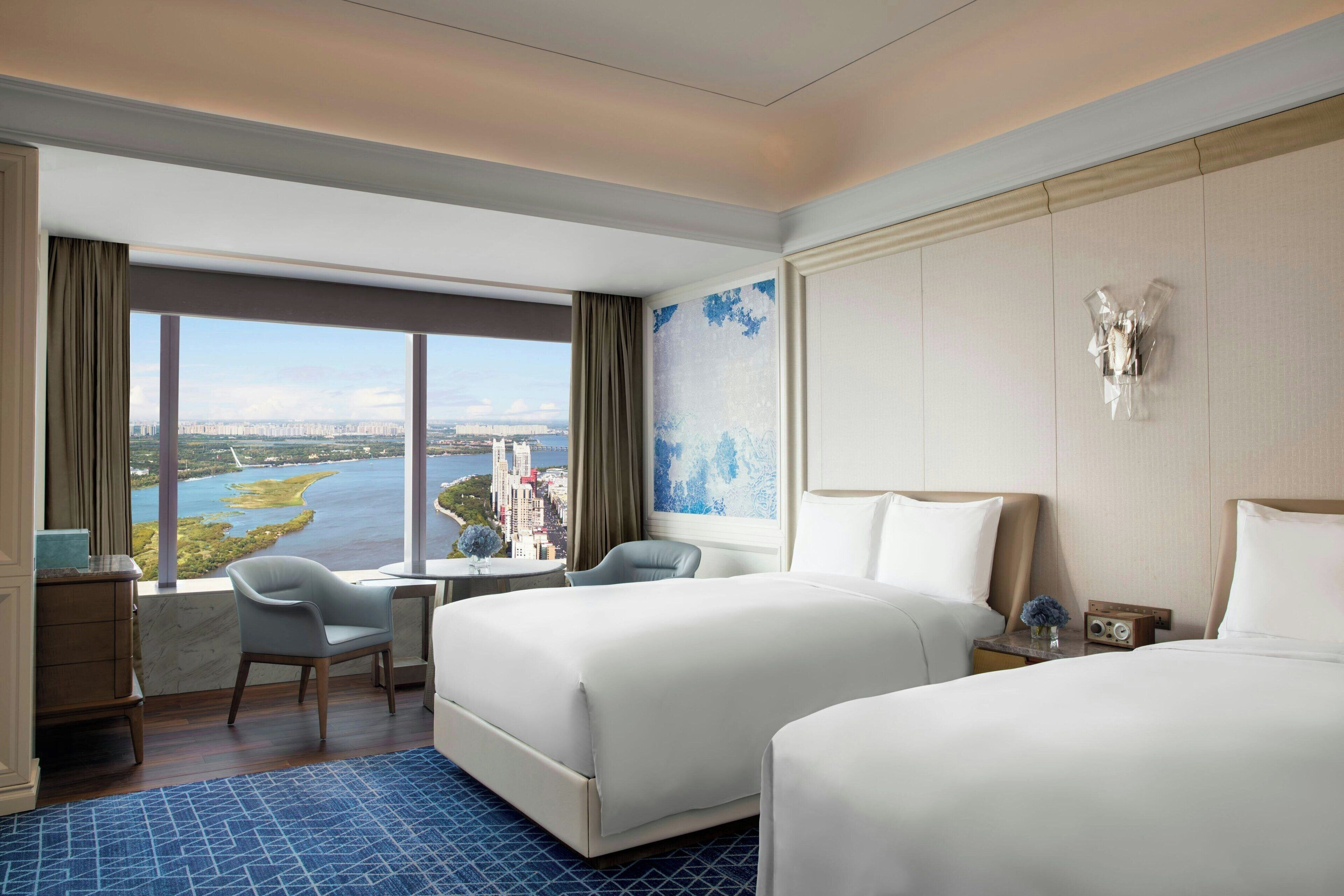 A hotel room with two beds, a desk, a chair, and a large window offering a city view.