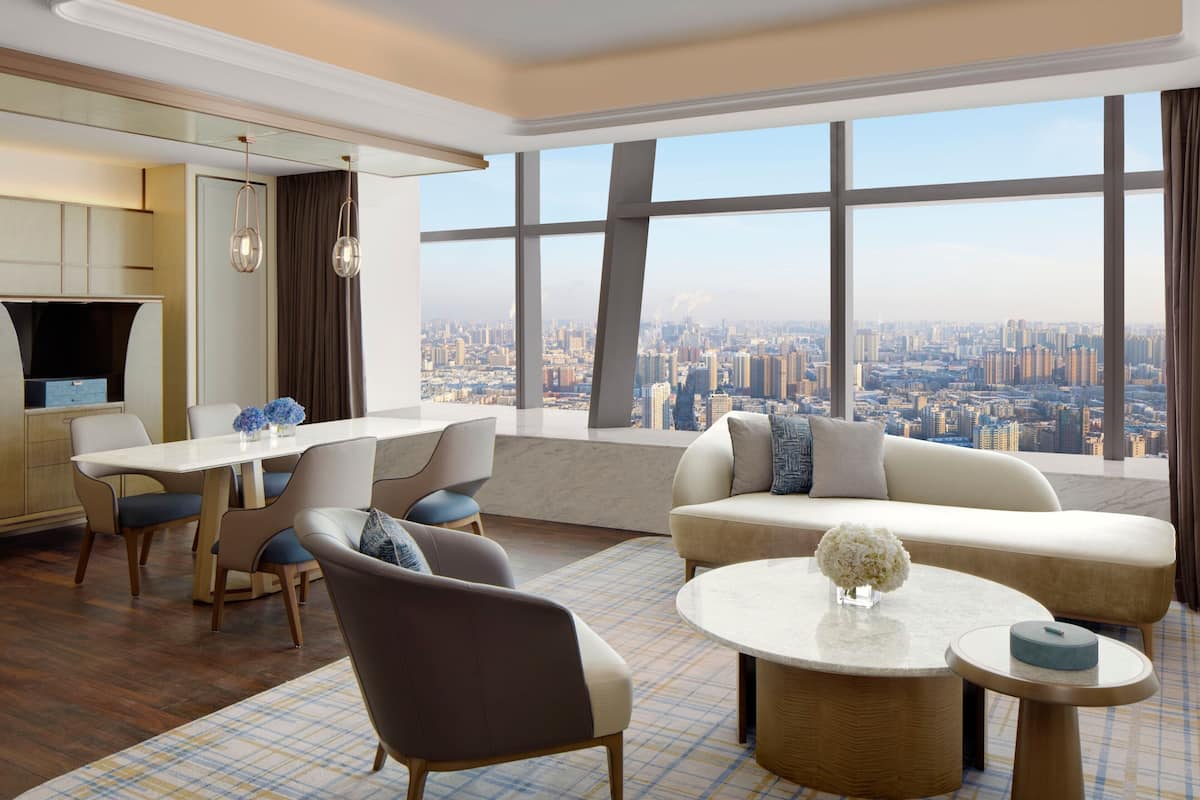 A modern hotel room with a dining area, a sofa, and a large window offering a city view.