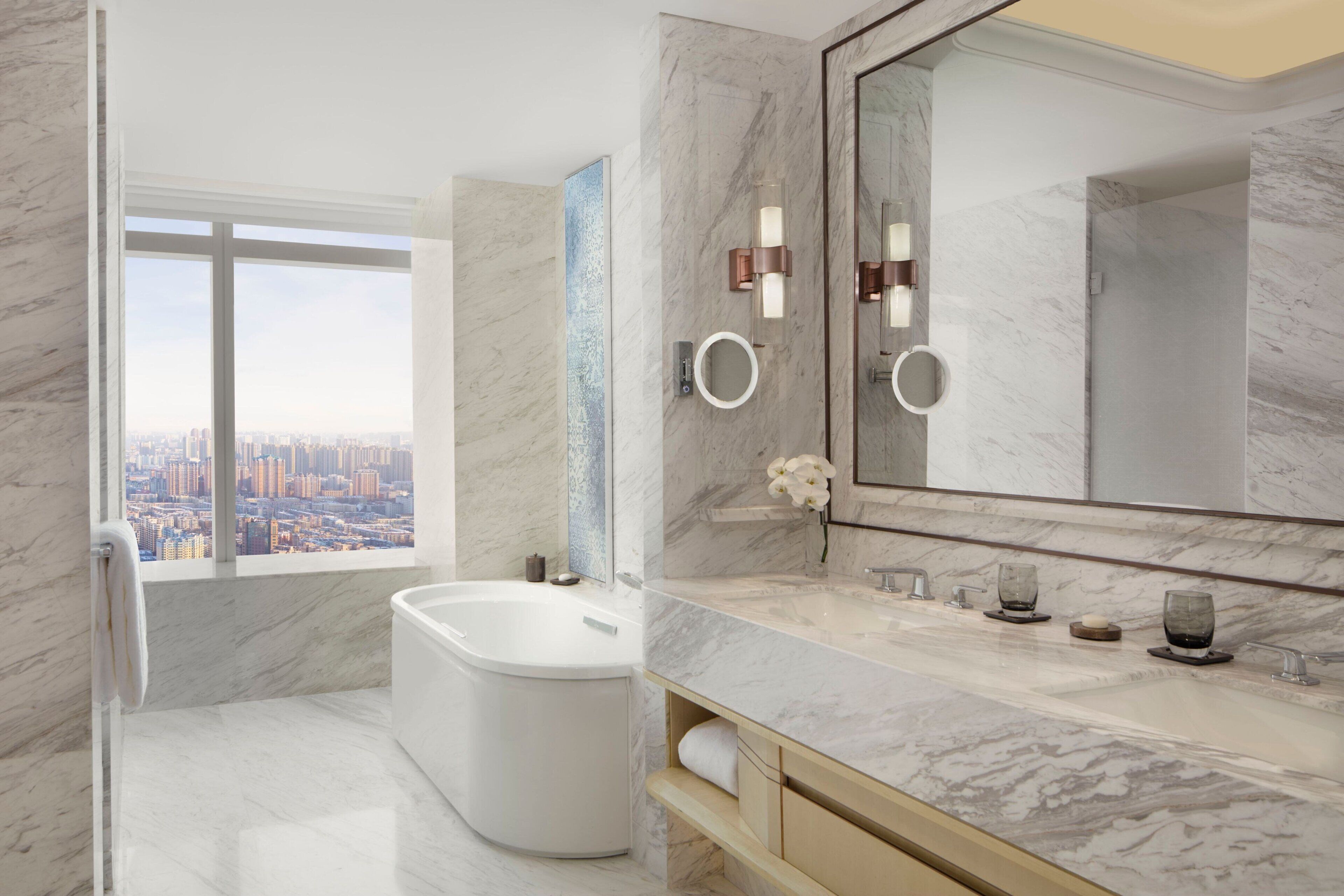 Un baño moderno con una bañera grande, un tocador con espejo y vistas a un paisaje urbano a través de un ventanal.