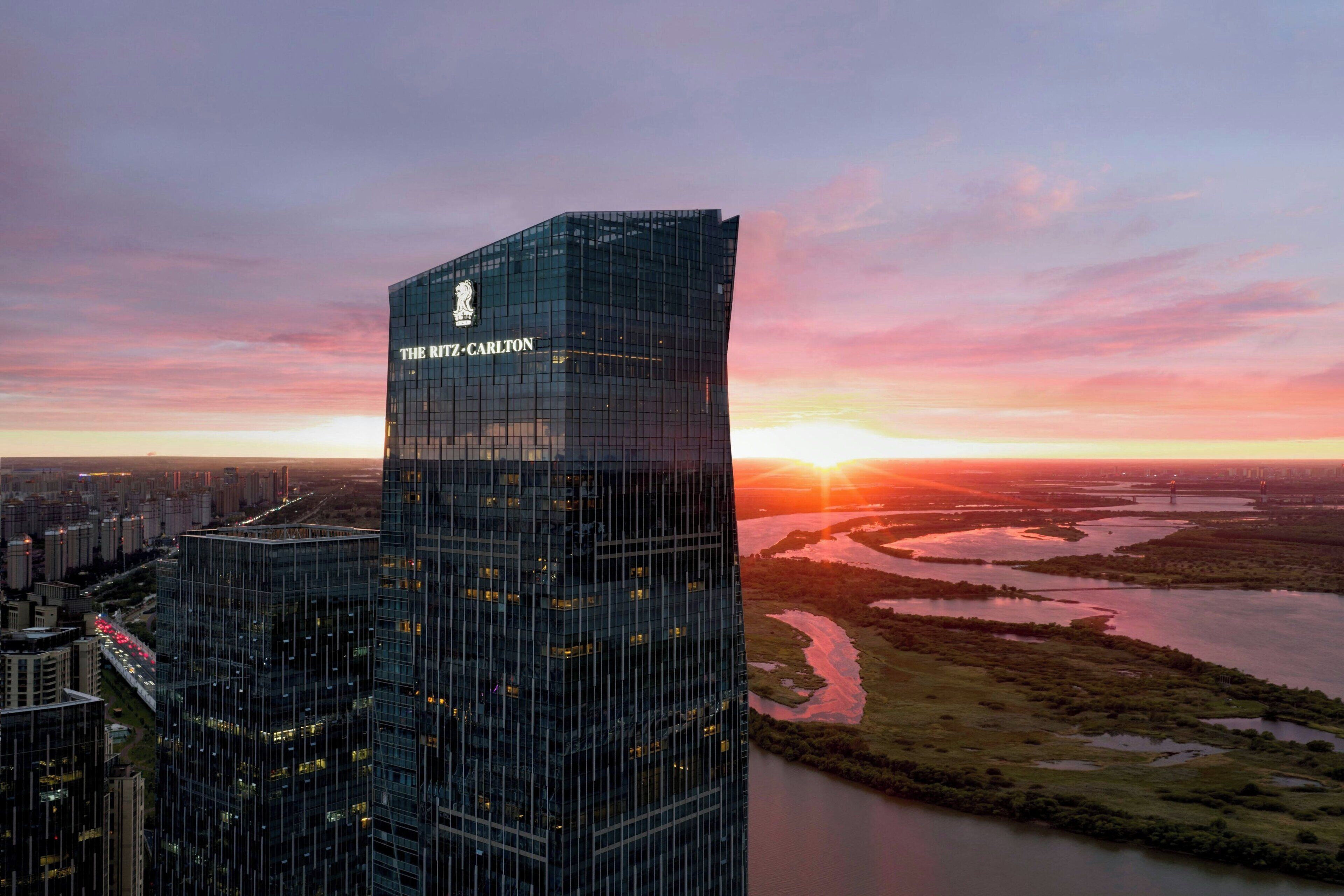 A high-rise building with "THE RITZ-CARLTON" signage, surrounded by a cityscape and a river at sunset.