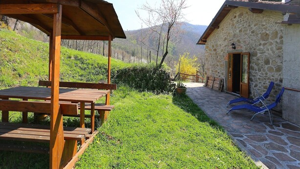 A wooden pergola with benches, a stone building with a door, and a stone pathway.