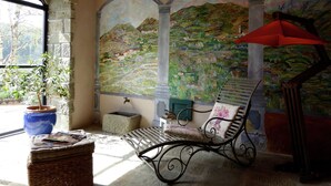 Una habitación acogedora con pared de piedra, un mural de un paisaje, una silla de mimbre, un paraguas rojo y una ventana con vistas a árboles.