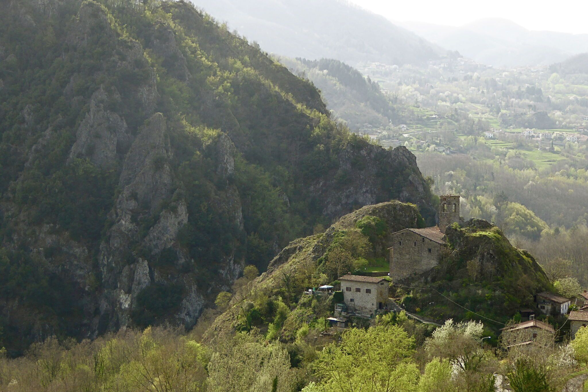 Paisaje montañoso con un edificio de piedra y una torre, rodeado de vegetación y un pueblo lejano.