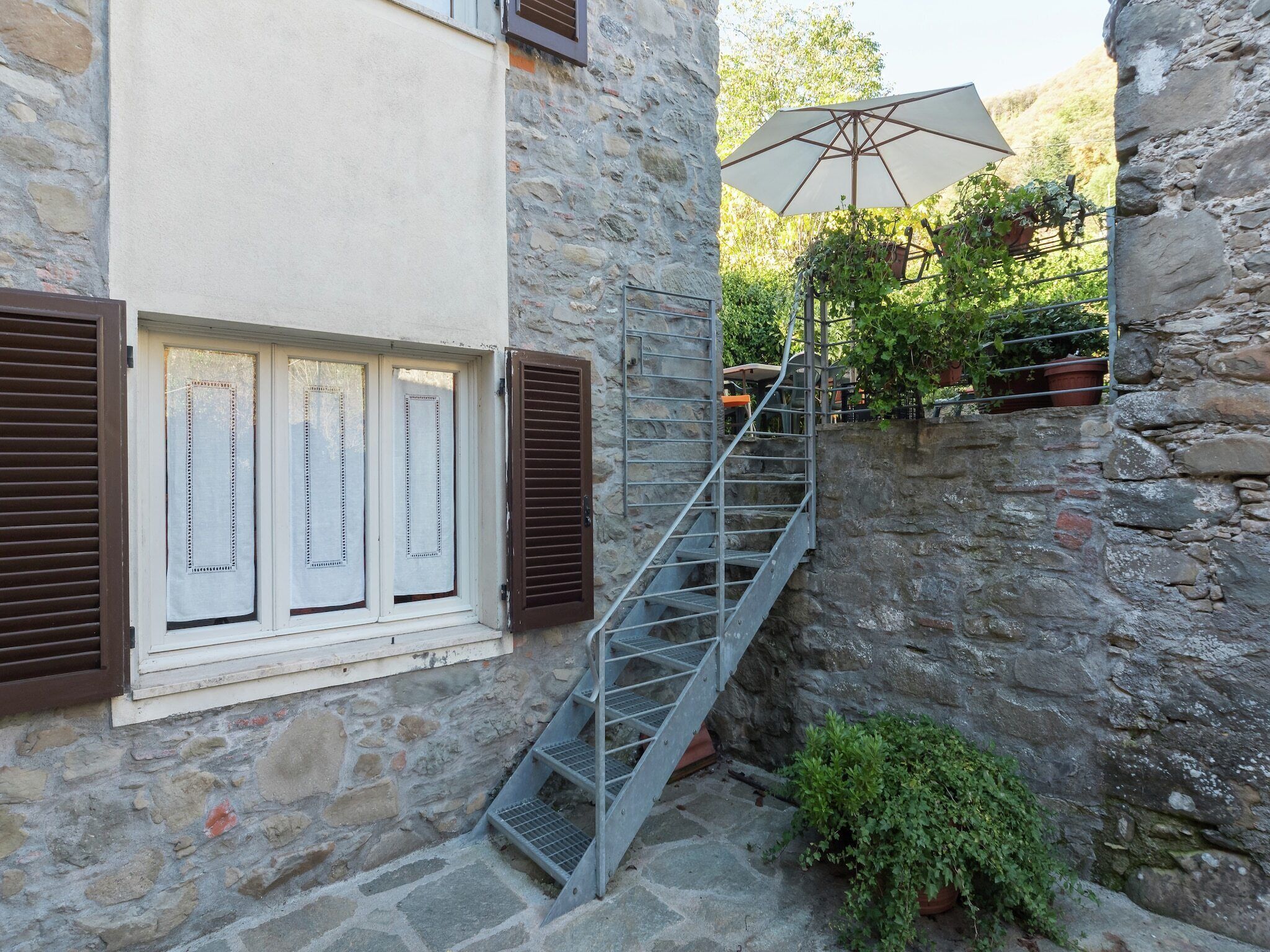 Un escalier en pierre menant à une fenêtre avec des volets, une rampe métallique et un pot de fleurs.