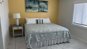 A bedroom with a bed, a nightstand, a lamp, a painting, and a window with blinds. - King Suite on Canal (Saint James City)