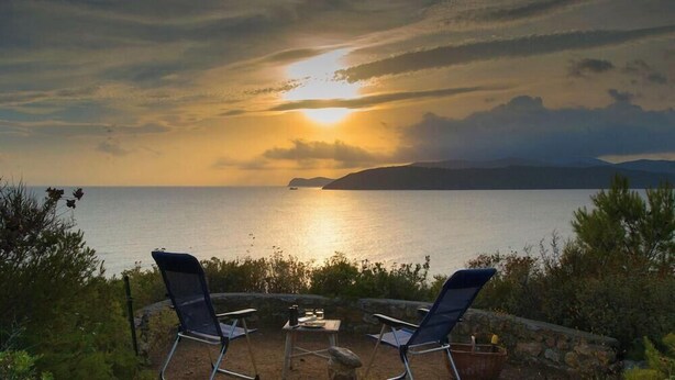 Dos sillones de relax mirando al atardecer sobre un cuerpo de agua, con un muro de piedra y follaje en primer plano.
