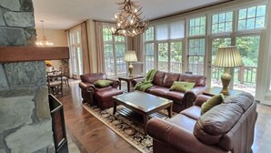 Ein geräumiges Wohnzimmer mit Ledersofas, einem Couchtisch und einem Kamin aus Stein. Große Fenster sorgen für natürliches Licht und Blick ins Grüne.