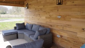 Una zona salotto esterna coperta con divano e poggiapiedi in vimini, pareti in legno e una lanterna.