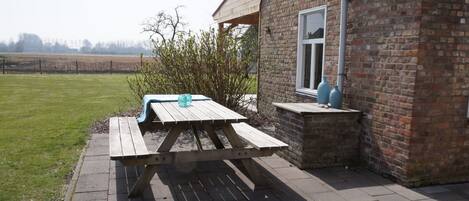 A wooden picnic table with a bench, a brick building, and a grassy field in the background.