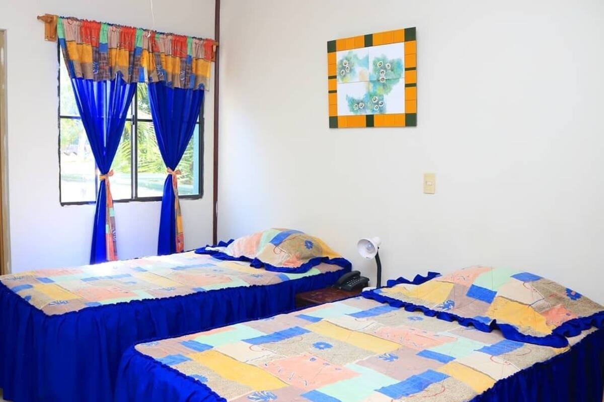 Ein Zimmer mit zwei Betten, jeweils mit einer blauen Bettdecke bezogen, und einem farbenfrohen Vorhang vor einem Fenster.