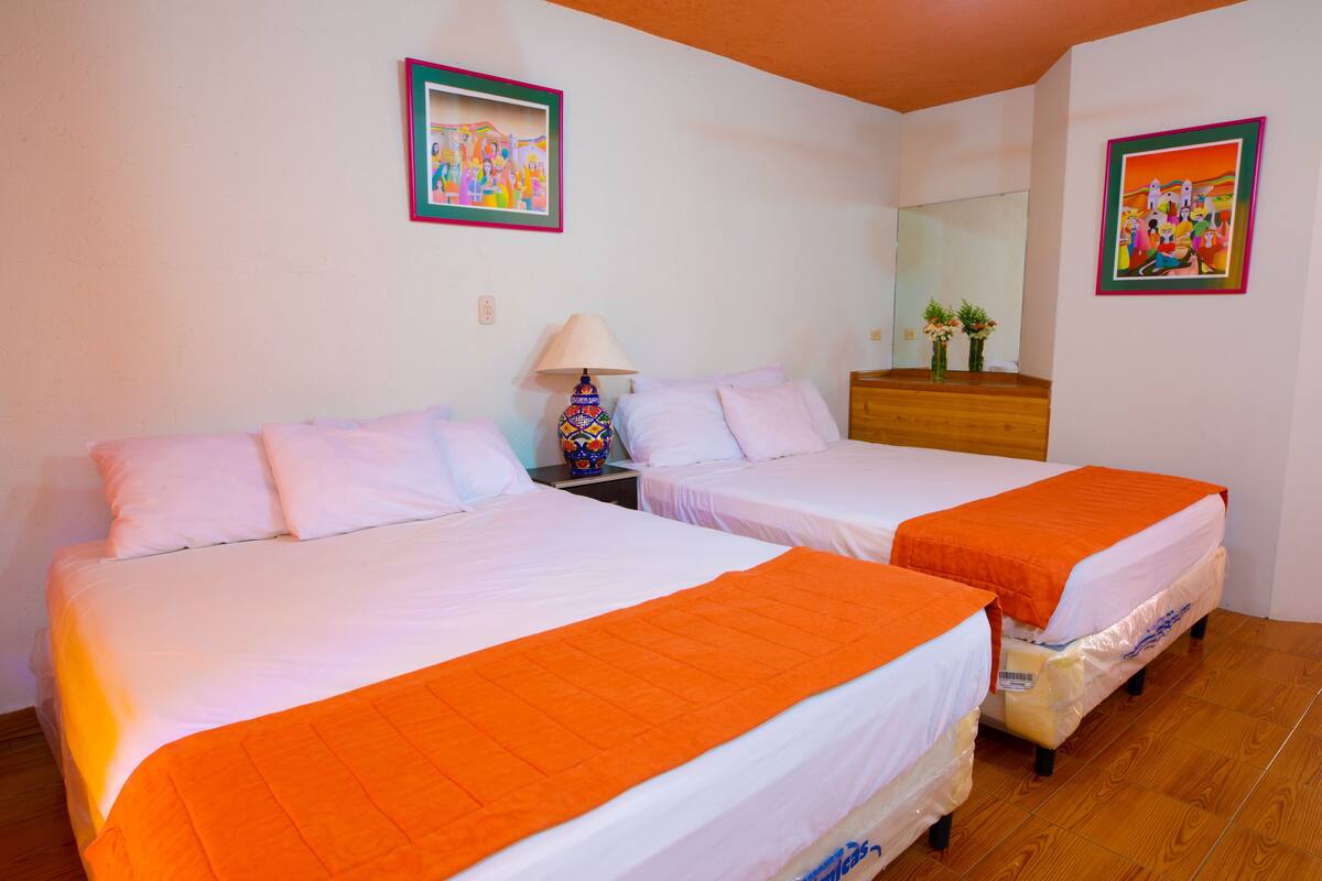 Ein Zimmer mit zwei Betten, jeweils mit weißer Bettwäsche und einer orangefarbenen Decke. An den Wänden hängen zwei Gemälde, ein Nachttisch mit einer Lampe und eine Holzkommode mit einem Spiegel.