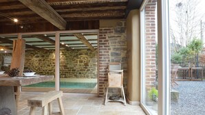 Un intérieur rustique avec des murs en pierre, un banc en bois, une petite table et une vue sur l’extérieur grâce à de grandes fenêtres.