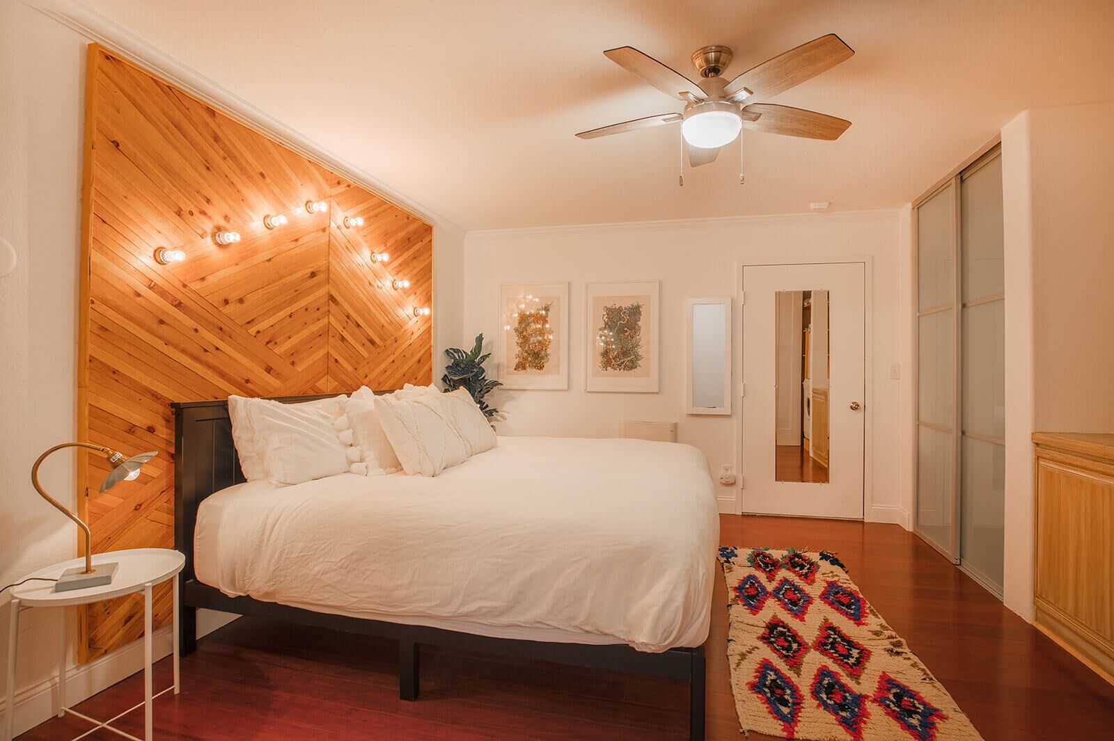 Ein Schlafzimmer mit Holz-Kopfteil, einem Bett mit weißer Bettwäsche, einem Deckenventilator, einer Nachttischlampe und einem gemusterten Teppich.