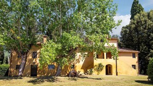 Una villa amarilla con un gran árbol en primer plano, un pequeño patio y un cielo despejado.