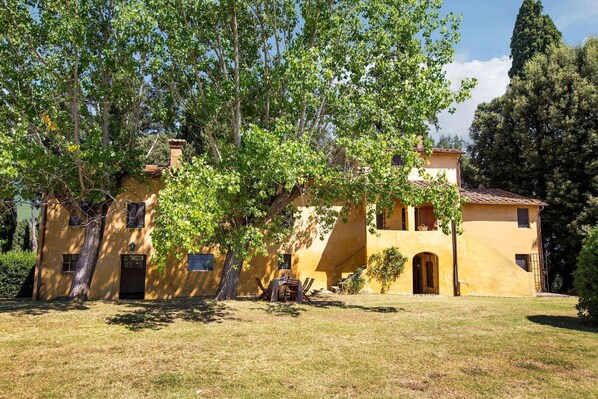 A yellow villa with a large tree in front, a small patio, and a clear sky.