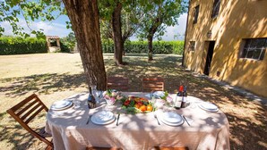 Mesa de comedor exterior con mantel blanco, sillas de madera y un centro de mesa con frutas.